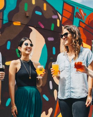 Four people are standing in front of a colorful mural, holding drinks, and smiling, creating a vibrant and cheerful atmosphere.