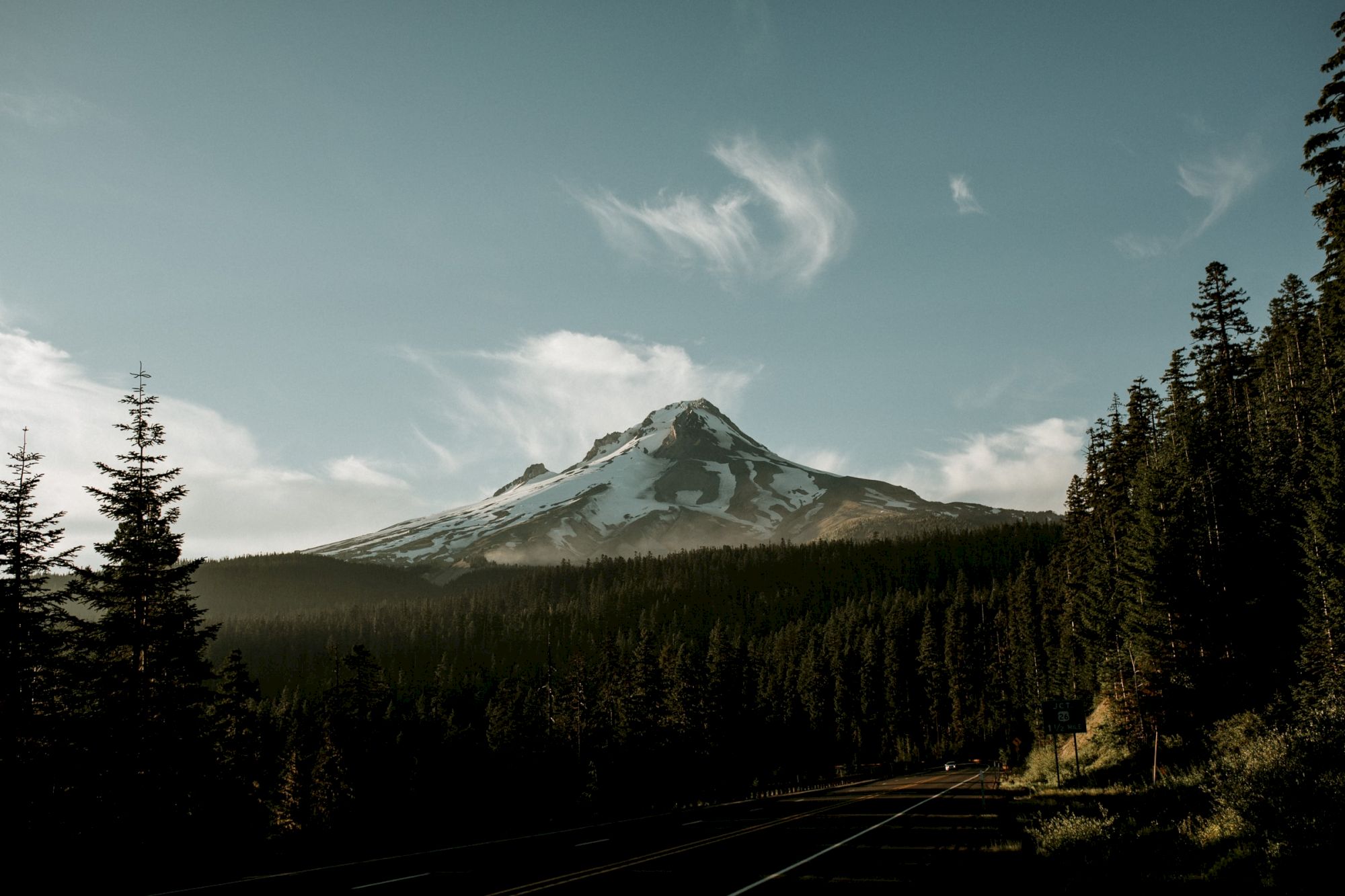 A snow-capped mountain stands tall under a clear sky with scattered clouds, surrounded by dense, evergreen forests and a road in the foreground.